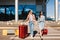 Two pretty girls in sunglasses happily walk along the pedestrian strip with red suitcases with airport on background