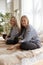 Two pretty twin sisters in black jeans and striped shirts with long hair sitting on sofa against background of window