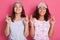 Two pretty women in pajamas and blind fold praying with crossed fingers, keep eyes closed, standing against pink wall, ladies with