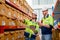 Two professional warehouse workers stand between shelves of products with one point to the box and discuss together about work in