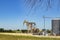 Two pump jacks at a pumping station with tanks and machinery with some farm buildings on the horizon under a blue sky with a few t