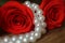 Between the two red roses lies a white pearl necklace on a wooden table brown. Macro.