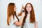 Two redheaded young women standing on isolated white backgroung, one curls her friend`s hair with styler, both look satisfied ,