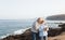 Two senior people standing on the cliff of the ocean and looking at cell. Hugging and smiling. Morning soon outdoor with clear sky