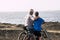 Two seniors at rocky beach with their bikes - couple of pensioners hugged together looking at the sea or ocean - healthy and