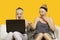 Two shocked young women using laptop sitting on sofa against yellow background