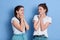 Two shy women friends european girls in casual clothes posing isolated over blue wall, ladies looking at each other keeping fists