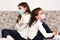 Two sisters sitting back to back on the sofa with medical protective face masks