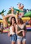 Two sisters, standing is front of attraction in theme park, holding popcorn in paper box, smiling, having fun. Pretty girls.