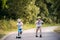 Two small boys with a helmet riding scooters on a road in park on a summer day.