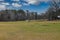 Two small brown horses grazing on green and yellow grass on the farm surrounded by bare winter trees and a sprinkler with blue sky