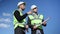 Two smart positive young men in hard hat and uniform gesturing high-five discussing building at background of clear blue