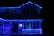 A two story house decorated with blue Christmas lights on the roofline and front porch with a lit Christmas tree in the window