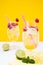 Two summer non-alcoholic cocktails with lime and raspberries stand on a white table on an yellow background.