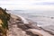 Two surfers walk up a rocky shoreline beach with surf rolling in and green hillside