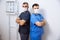 Two surgeons in sterile uniforms standing back to with arms folded and looking camera on white background