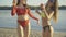 Two tanned slim women clinking bottles and dancing on sandy summer beach. Middle shot portrait of joyful young Caucasian