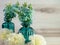 Two teal glass vases up close with blue hydrangeas flowers and white carnations in front of them on a beachwood background.  Home