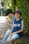 Two teenage brothers one of them looking confident in the camera sitting on a stone wall under trees in a park in summer holidays