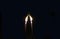 Two thin candles stretch their lights towards each other. The candles are in complete darkness.