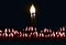 Two thin candles stretch their lights towards each other. Their fire merges into one flame. The candles are in complete darkness.