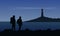 Two tourist with backpacks standing on the sea shore and watching the shining lighthouse on the island under blue night sky