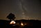 Two tourists at burning campfire in front of tent, photo camera on tripod under dark starry sky.