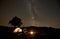 Two tourists at burning campfire in front of tent, photo camera on tripod under dark starry sky.