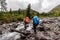 Two tourists with large backpacks cross the river in rubber boots.