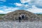Two tourists looking at ruins of the architecturally significant Mesoamerican pyramids and green grassland located at at