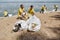 Two trash bags on the sand, group of young eco activists or volunteers in uniform collecting plastic waste and rubbish