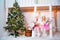 Two twin sisters in doggie dresses at the front of a white wooden house are playing. Girls and poodles decorated for Christmas and