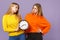 Two upset young blonde twins sisters girls in vivid colorful clothes holding round clock isolated on pastel violet blue
