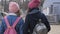 Two walking girls, teenagers in caps, coats, jackets with backpacks, back view