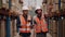 Two warehouse workers wearing hardhats and reflective jackets maintain the detail of manufactured goods in the warehouse