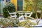 Two white Adirondack chairs with a table between them in the snow in front of a colorful house with green shutters and shrubbery