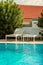 Two white beach chairs next to a pool with warm blue water. Hotel pool setting. Big green bush plant and roof of a house in the