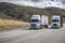 Two white big rigs semi trucks with different loaded semi trailers climbing uphill on the mountain road in California