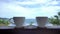 Two white coffee cups are placed over a wooden fence with a beautiful view of the sea close up