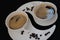 Two white cups with black coffee are standing on two white plates with coffee beans against black background