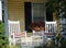 Two white rocking chairs are on the front porch of a yellow house