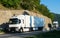 Two white trucks carrying the goods, going on a beautiful road near the mountain