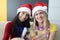 Two women friends in santa hats are sitting on sofa at home and drinking champagne