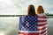 Two women friends with USA national flag on their shoulders standing together outdoors on lake shore. Patriotic girls celebrating