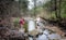 Two women hiking in the woods across a water stream.