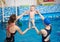 Two women holding happy laughing baby doing exercising over water in air during swimming classes. Baby relaxing in pool