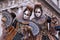 Two women in masks and costume, with decorated fans, standing in front of the arches at St Marks Square during the Venice Carnival