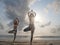 Two women practice tree yoga asana at sea beach in sunlight