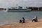 Two women sitting together on a pebble beach in the summer with a ferry passing