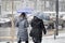 Two women walking under umbrellas in city blizzard and traffic behind them